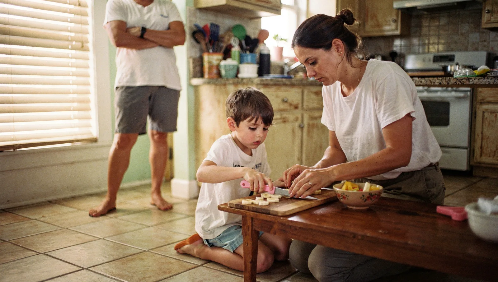 A therapist gently guides a young boy's hands to slice a banana with a plastic knife in a sunlit kitchen.