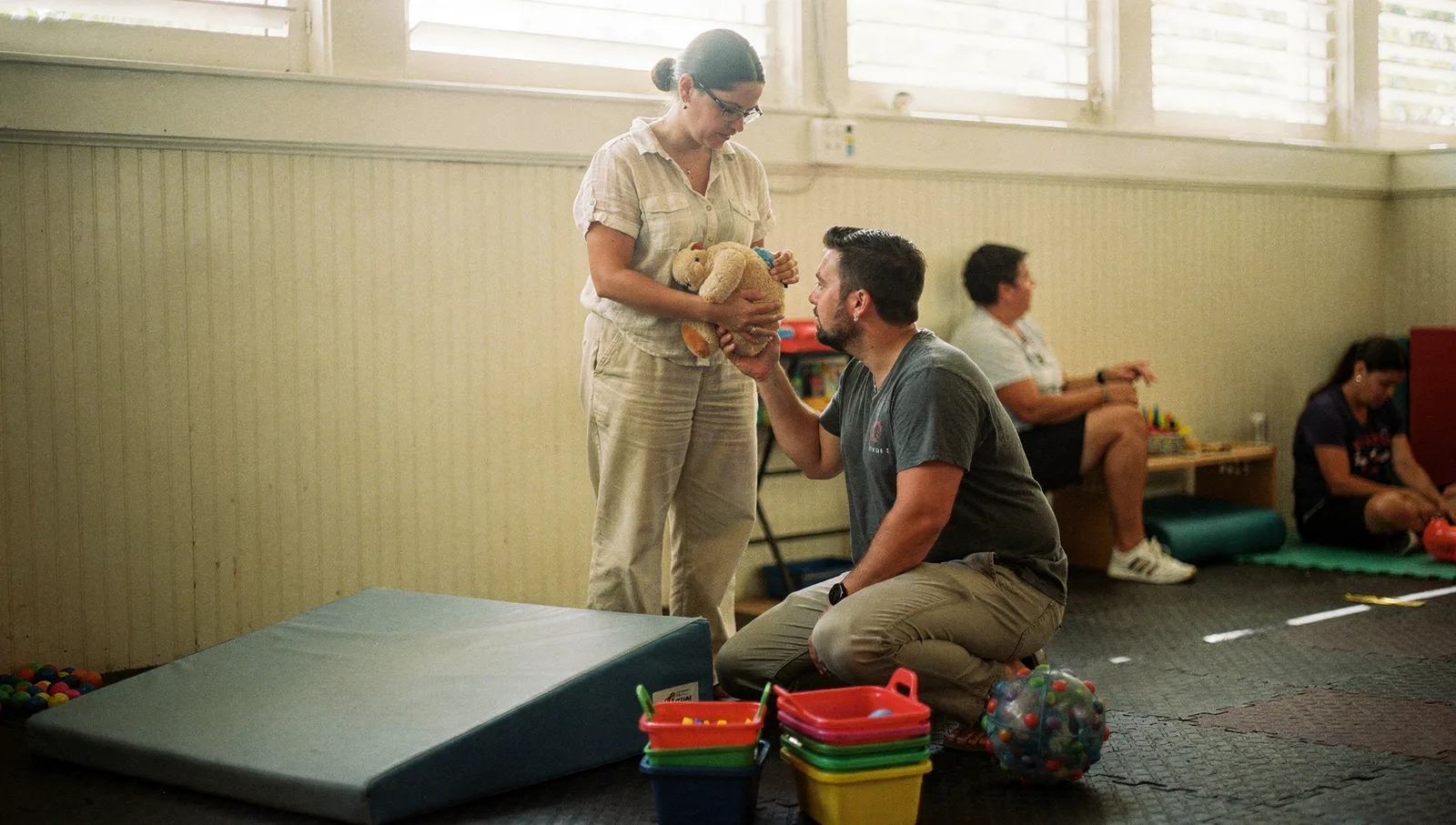 A behavior analyst demonstrates prompting techniques with a plush toy to a trainee in a sunlit sensory gym room.