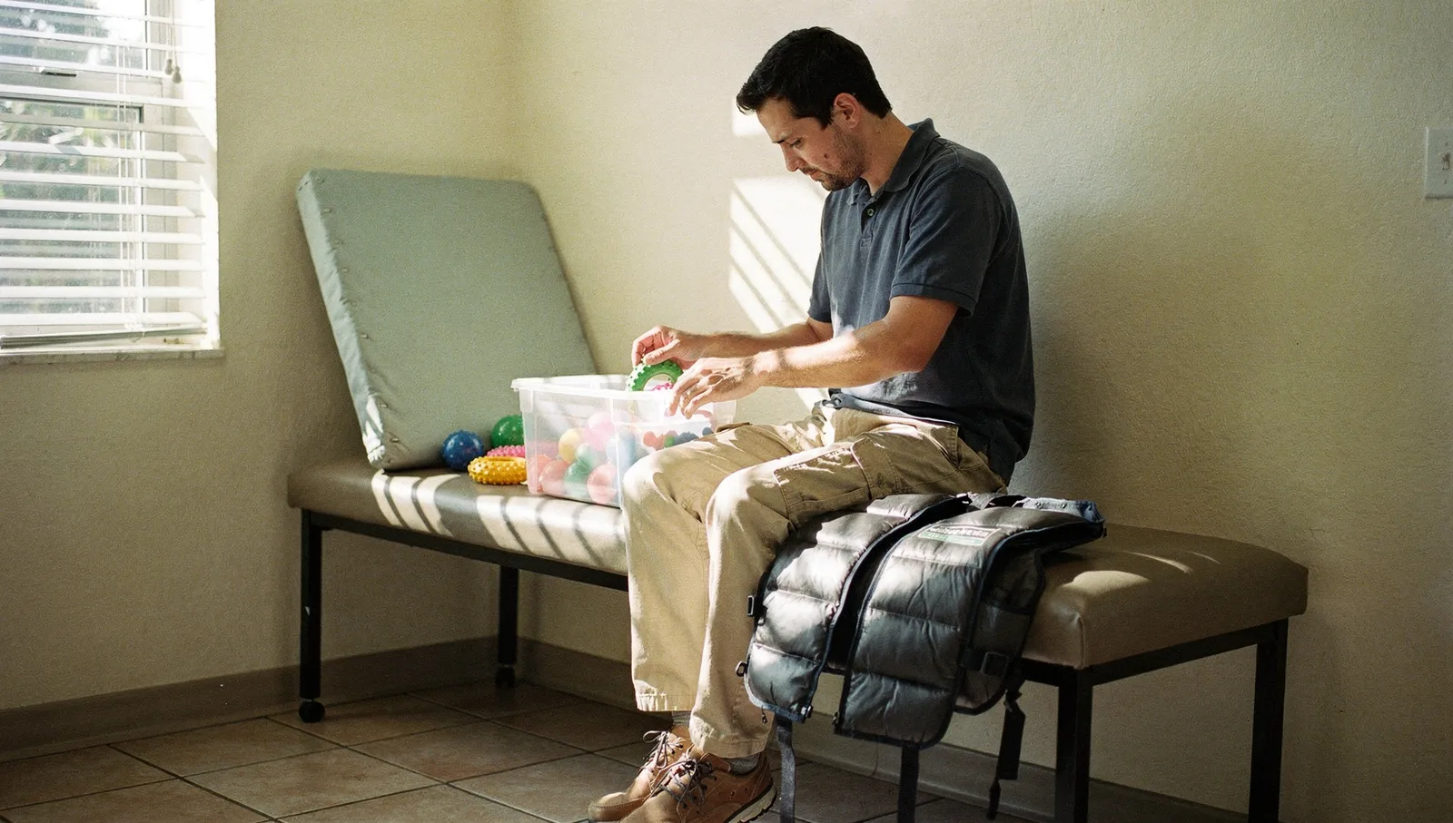 A clinical technician arranges sensory toys in plastic bins in a pediatric therapy room beside a padded bench.