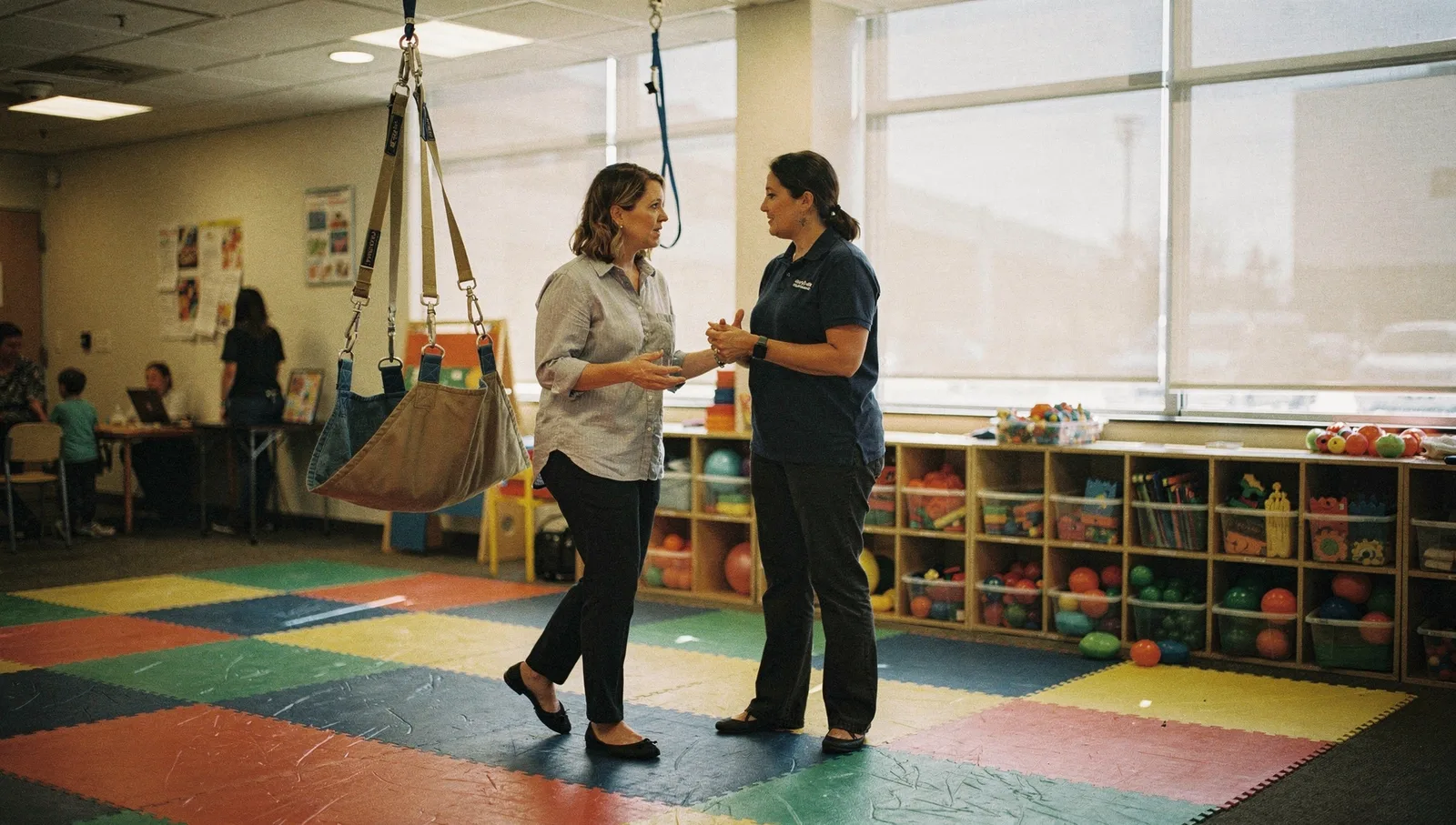 Two staff members talk near a sensory swing and cubbies on foam floor tiles inside a pediatric ABA therapy clinic.