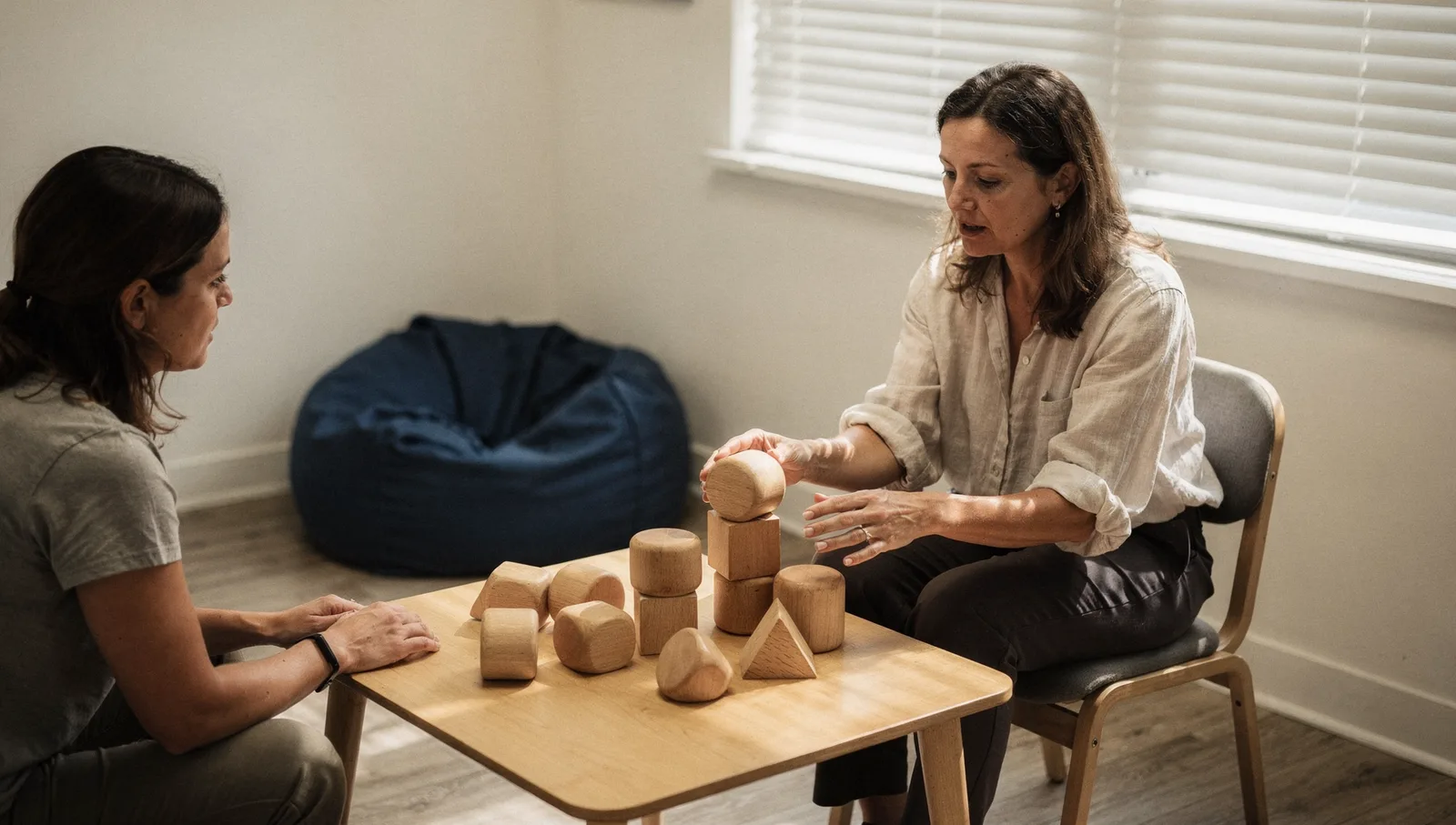 Therapist demonstrating a play technique with wooden blocks on a low table to an attentive mother in a bright clinic.