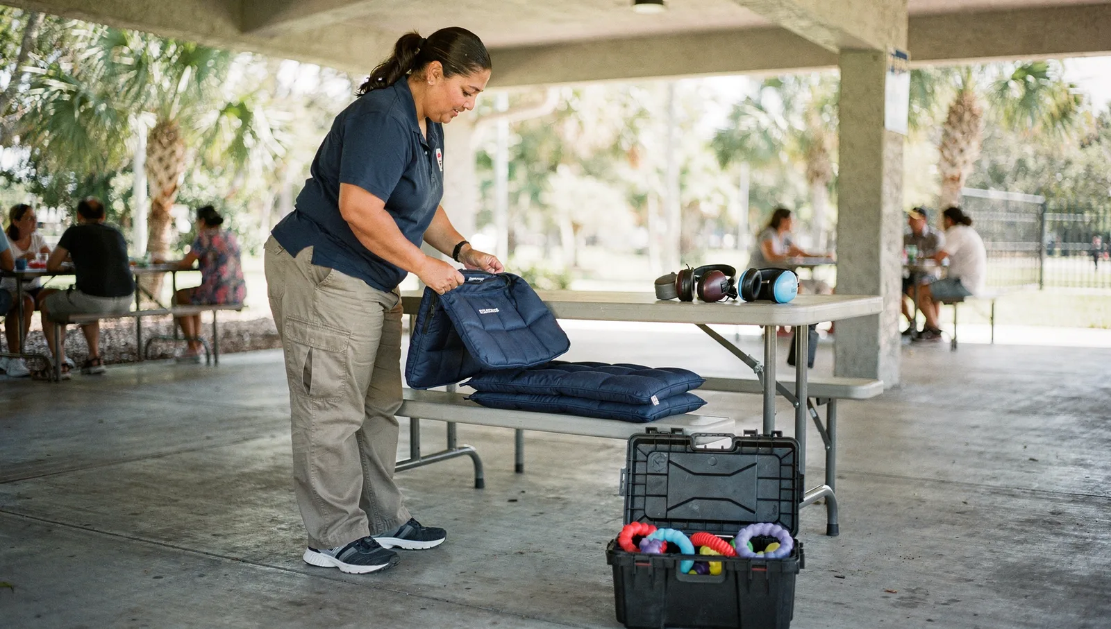 A woman arranges sensory quiet-space equipment on a bench inside a shaded park pavilion surrounded by palm trees.
