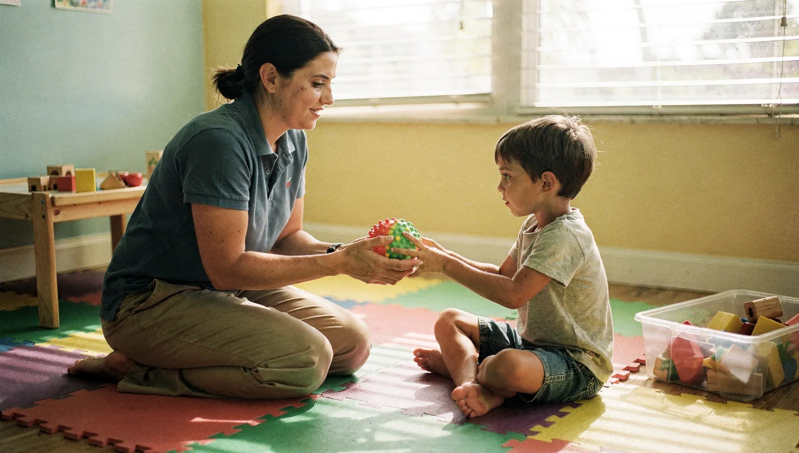 A female therapist sits on a foam mat offering a textured sensory ball to a young boy in a sunlit therapy room.