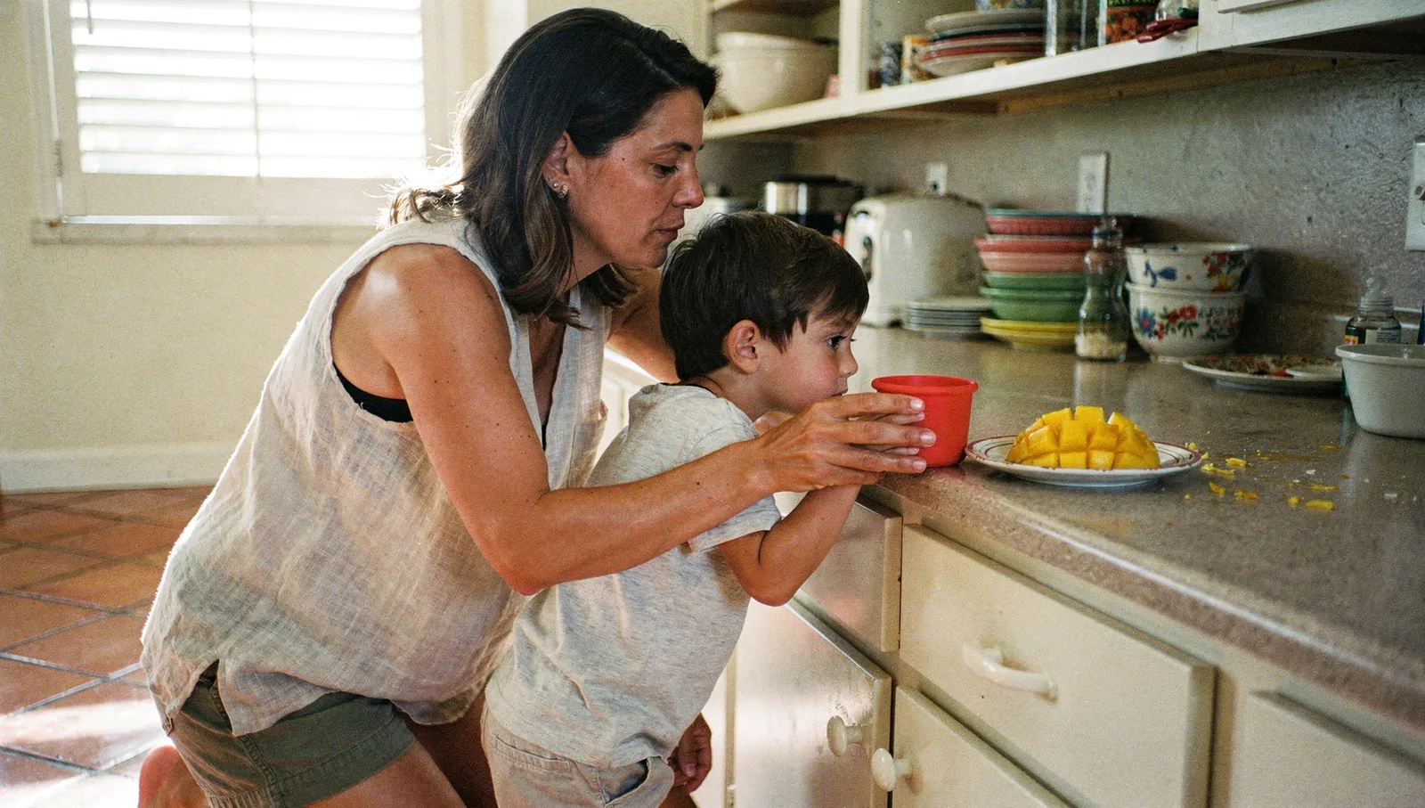 A mother kneels beside her young son in a kitchen, holding a cup just out of reach as he gestures toward it.